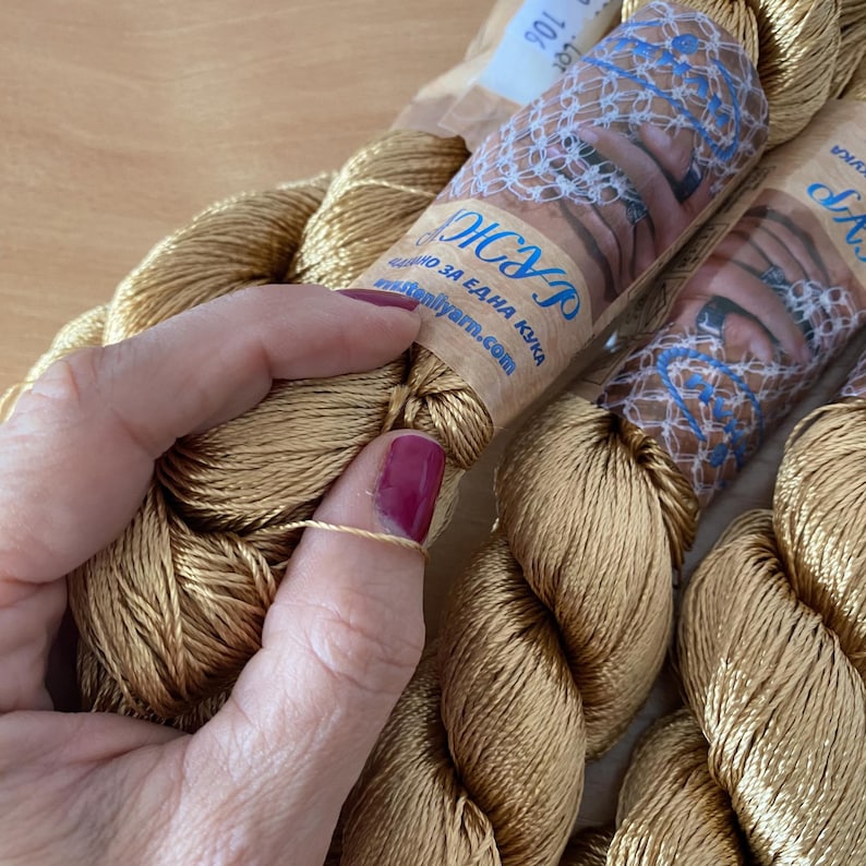 K&ouml;nnte beinhalten: Nahaufnahme mehrerer Str&auml;nge goldfarbenen Stickgarns. Das Garn ist eng gewickelt und hat einen leichten Glanz. Das Etikett auf dem Strang enth&auml;lt Text in einer Fremdsprache. Eine Hand mit bordeauxfarbenem Nagellack h&auml;lt einen Faden des Garns.