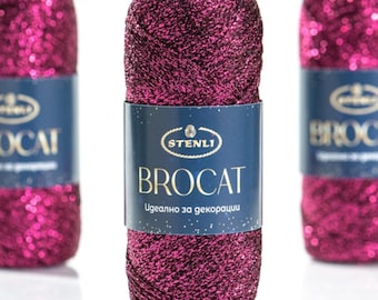Può includere: Tre matasse cilindriche di filato metallico color magenta scuro e nero. Ogni matassa presenta un'etichetta di carta blu con il testo STENLI BROCAT e altro testo in cirillico. Il filato ha un aspetto scintillante e testurizzato con fibre metalliche intrecciate.