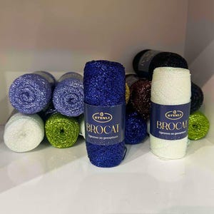 May include: A collection of Stenli Brocat yarn skeins in various colours including blue, white, green, and dark brown. Each cylindrical skein features a dark blue paper label with the text Stenli Brocat and metallic fibres.