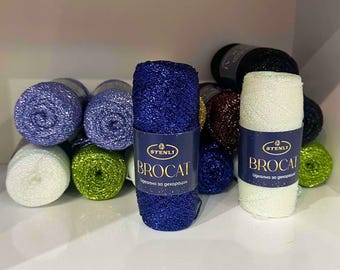 May include: A collection of Stenli Brocat yarn skeins in various colors including blue, white, green, and dark brown. Each cylindrical skein features a dark blue paper label with the text Stenli Brocat and metallic fibers.