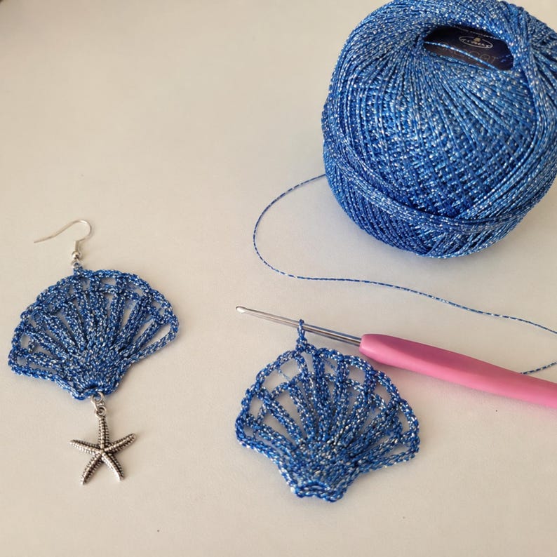 May include: A pair of blue and silver metallic crochet shell earrings, one attached to a silver starfish charm and a hook, the other attached to a pink crochet hook, accompanied by a matching ball of blue and silver metallic crochet thread.
