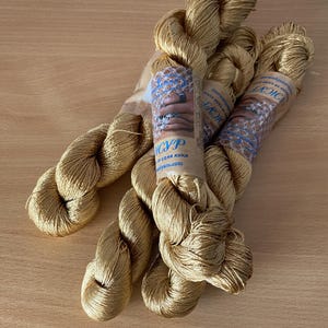 K&ouml;nnte beinhalten: Drei Str&auml;nge goldfarbenes Garn liegen auf einer hellbraunen Holzoberfl&auml;che. Jeder Strang ist mit einem Papieretikett mit Text und einem Handbild umwickelt. Das Garn hat ein weiches, gl&auml;nzendes Aussehen.
