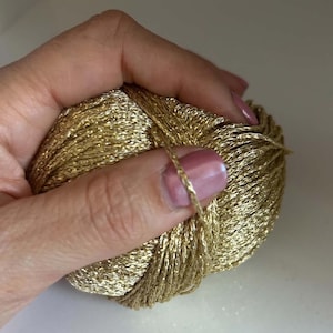 Puede incluir: Un ovillo de hilo dorado metálico con una textura brillante y reflectante. El hilo está enrollado en una forma compacta y redondeada, mostrando un acabado luminoso que capta la luz a través de sus finas hebras de fibra sintética.