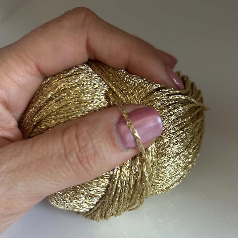 May include: A ball of metallic gold yarn with a shimmering, reflective texture. The yarn is wound into a compact, rounded shape, showcasing a bright, reflective finish that catches the light across its thin, twisted strands of synthetic fibre.