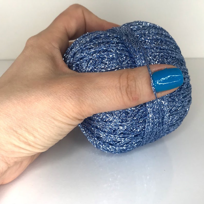 May include: A ball of metallic blue yarn with a shimmering, reflective texture. The yarn is wound into a compact, rounded shape, showcasing a bright, cool-toned blue color with silver highlights throughout the twisted fibers of the thread.