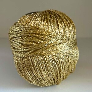 May include: A spherical ball of metallic gold yarn featuring a twisted, shimmering texture. The thread is wound tightly in a continuous pattern, creating a reflective surface that catches the light across the entire rounded form of the yarn.