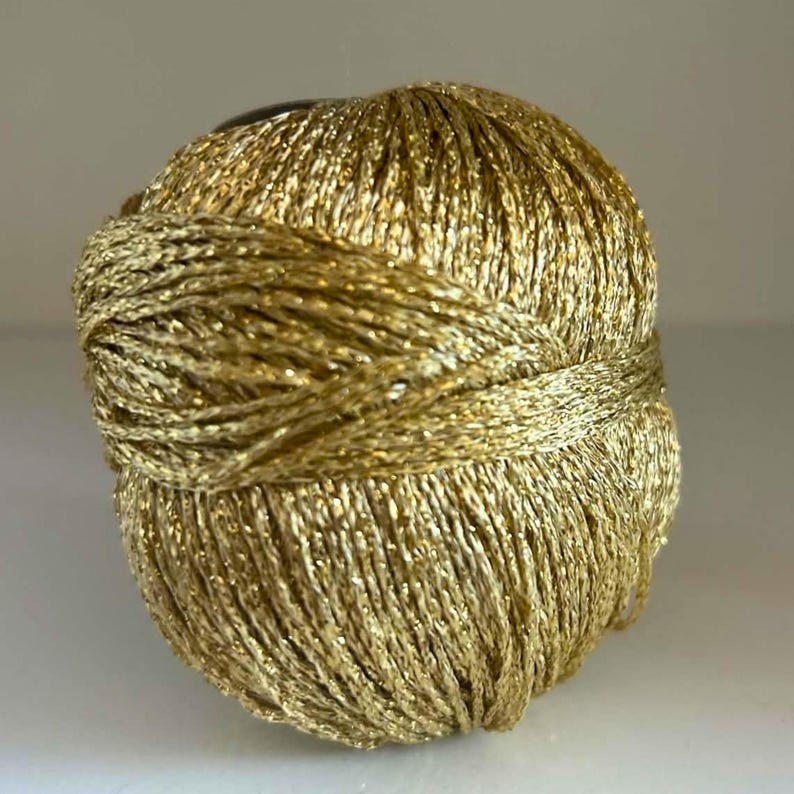 May include: A spherical ball of metallic gold yarn featuring a twisted, shimmering texture. The thread is wound tightly in a continuous pattern, creating a reflective surface that catches the light across the entire rounded form of the yarn.