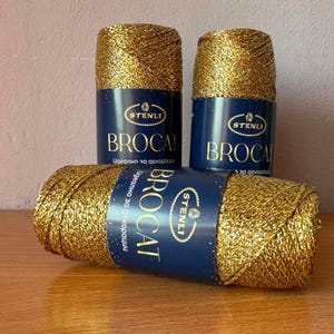 May include: Three skeins of metallic gold yarn, each wrapped in a dark blue paper label. The labels feature the brand name STENLI in an oval logo, the word BROCAT in large gold letters, and additional text in a smaller white font.
