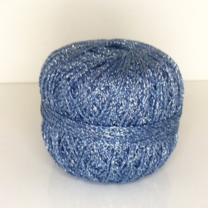May include: A round ball of yarn featuring a vibrant blue color intertwined with shimmering silver metallic threads. The yarn is wound into a compact, dense sphere with a visible texture created by the combination of blue and silver fibers.