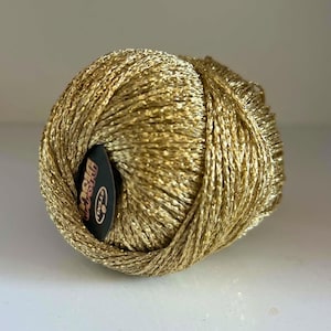 May include: A spherical ball of metallic gold yarn with a shimmering, reflective texture. A small, circular black label is tucked into the side of the yarn ball, featuring the white text "STENLI" and the word "Disco" printed in a stylised font.