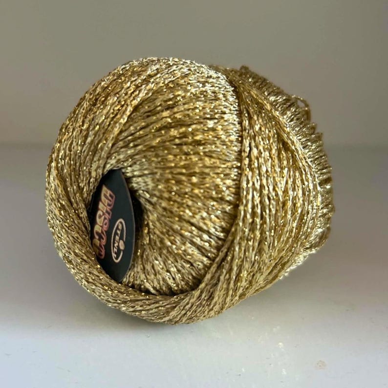 May include: A spherical ball of metallic gold yarn with a shimmering, reflective texture. A small, circular black label is tucked into the side of the yarn ball, featuring the white text "STENLI" and the word "Disco" printed in a stylised font.