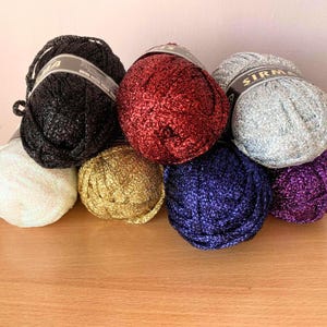 May include: A selection of colourful yarn skeins in shades of black, red, gold, silver, blue, white, and purple. The yarn has a metallic sheen, suitable for decorative knitting or crochet. The skeins are arranged on a wooden surface, likely for crafting projects.