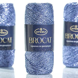 May include: A skein of metallic blue yarn with a shimmering texture. The center features a dark blue paper label with the gold text STENLI BROCAT and additional white text. The yarn is wound into a cylindrical shape with a visible cross-hatch pattern.