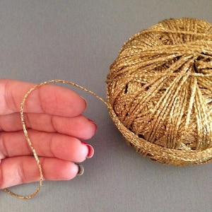 May include: A spherical ball of metallic gold yarn with a shimmering, reflective texture. A single strand of the thin, twisted gold thread extends from the main ball, showcasing the consistent metallic finish and the fine, cord-like construction.