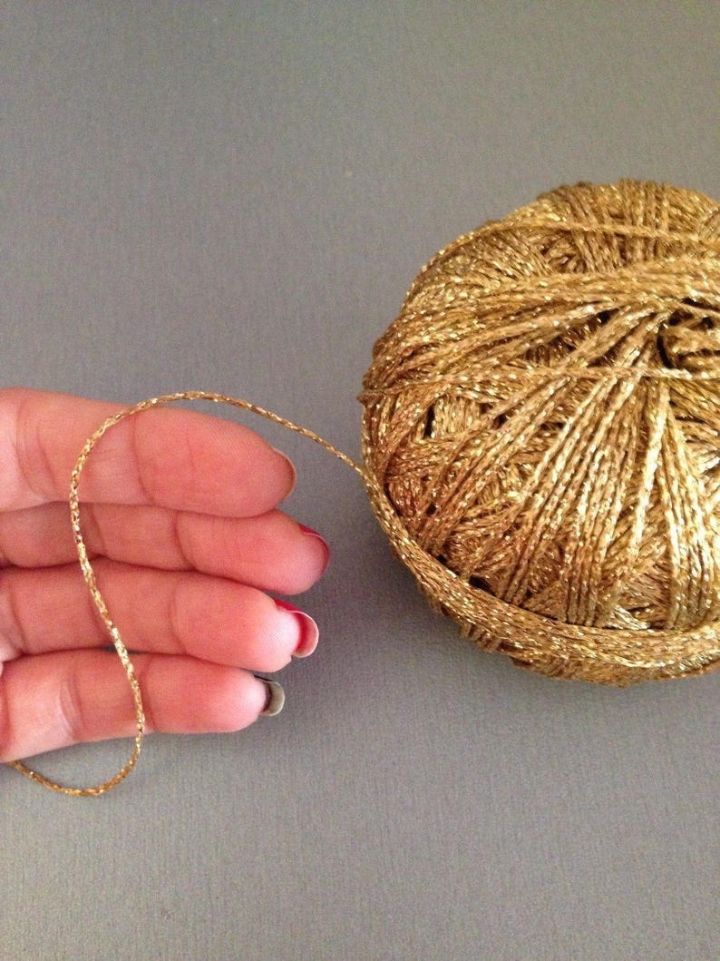 May include: A spherical ball of metallic gold yarn with a shimmering, reflective texture. A single strand of the thin, twisted gold thread extends from the main ball, showcasing the consistent metallic finish and the fine, cord-like construction.