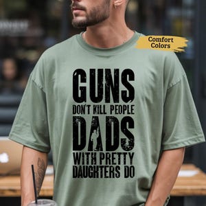 May include: Sage green t-shirt with the text "GUNS DON'T KILL PEOPLE DADS WITH PRETTY DAUGHTERS DO" in black. The shirt has a relaxed fit and short sleeves. The words "Comfort Colors" are printed on the shirt.