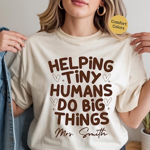 May include: A beige t-shirt featuring the brown text "HELPING TINY HUMANS DO BIG THINGS", with hearts and the name "Mrs. Smith". The shirt is a comfort color and is being worn.