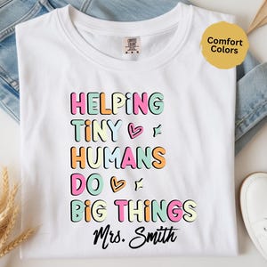 May include: White t-shirt featuring the phrase "HELPING TINY HUMANS DO BIG THINGS Mrs. Smith" in a colourful, bubble letter font. The text is in pink, green, orange, and yellow. The t-shirt is laid on blue denim jeans and next to white trainers.