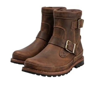 Puede incluir: Un par de botines de cuero marrón con punta redondeada, suela gruesa de tacos negra y dos correas ajustables con hebillas de metal en tono latón antiguo. Las botas presentan costuras visibles en el mismo tono y un cuello acolchado superior.