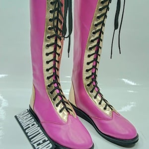 Puede incluir: Un par de botas altas rosas con detalles dorados y cordones negros. Las botas tienen suela negra y ribete dorado en la parte superior. Las botas son de material liso y tienen un diseño clásico. El texto "BABAR FOOTWEAR" es visible.