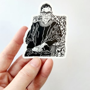 May include: Black and white illustration of a woman wearing a judicial robe and glasses, sitting in a chair with a patterned back. The illustration is printed on a white sticker.