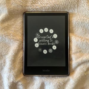 May include: A black Kindle e-reader with a dark screen showing the text "introverted & willing to discuss books" encircled by daisy graphics. The e-reader rests on a white, textured surface.