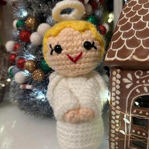May include: A handmade crochet angel figurine with a halo, blonde hair, and a white robe. The angel has black eyes and a red smile. A Christmas tree and a gingerbread house are in the background.