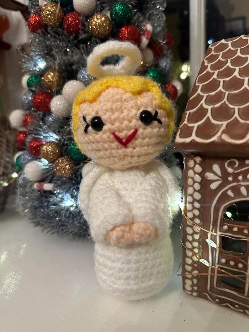 May include: A handmade crochet angel figurine with a halo, blonde hair, and a white robe. The angel has black eyes and a red smile. A Christmas tree and a gingerbread house are in the background.