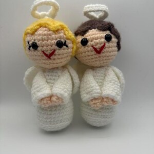 May include: Two handmade crochet angel dolls. One angel has blonde hair, the other has brown hair. Both have white wings, halos, and smiling red mouths. The dolls are made of a soft, textured yarn.