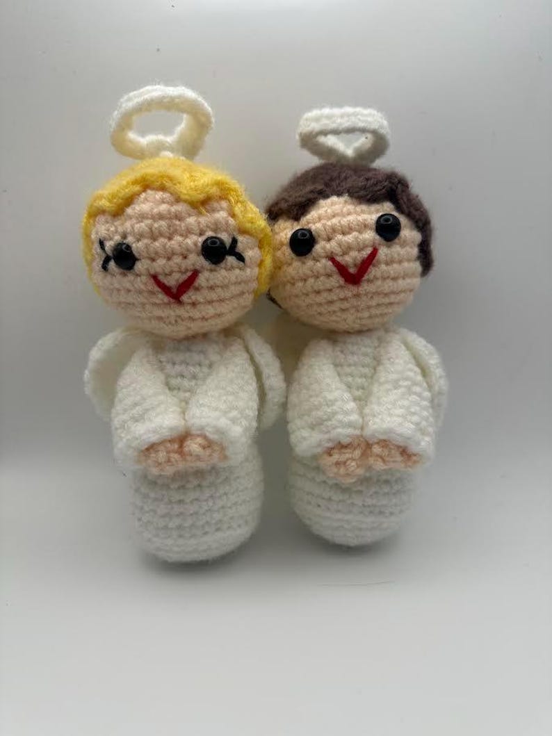 May include: Two handmade crochet angel dolls. One angel has blonde hair, the other has brown hair. Both have white wings, halos, and smiling red mouths. The dolls are made of a soft, textured yarn.