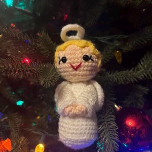 May include: A handmade crochet angel Christmas ornament. The angel has a yellow head, a white halo, and white robes. The angel's face has black eyes and a red smile. The ornament is hanging on a Christmas tree with colorful lights and ornaments.