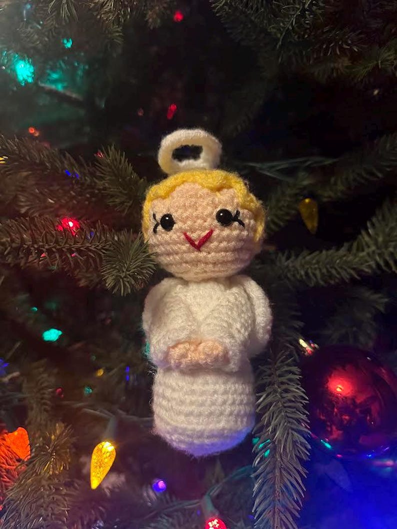 May include: A handmade crochet angel Christmas ornament. The angel has a yellow head, a white halo, and white robes. The angel's face has black eyes and a red smile. The ornament is hanging on a Christmas tree with colorful lights and ornaments.