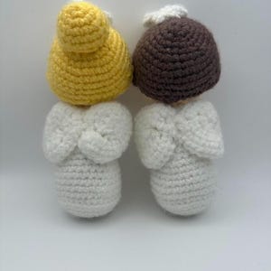May include: Two handmade crochet angel dolls. One doll has a yellow head and the other has a brown head. Both dolls have white bodies and wings. The dolls are made of yarn and are on a white background.
