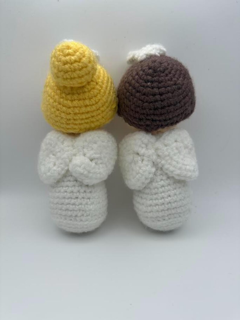 May include: Two handmade crochet angel dolls. One doll has a yellow head and the other has a brown head. Both dolls have white bodies and wings. The dolls are made of yarn and are on a white background.