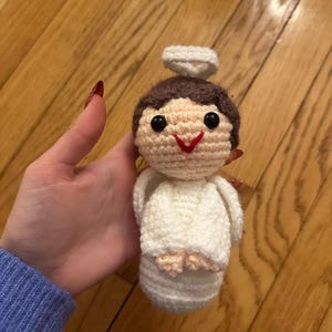 May include: A handmade crochet angel doll with a halo. The doll has a light brown head, black eyes, and a red stitched smile. The angel is wearing a white robe and has light pink feet. The doll is approximately 4 inches tall.