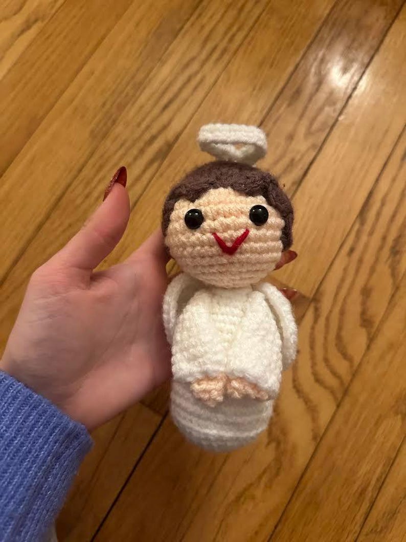 May include: A handmade crochet angel doll with a halo. The doll has a light brown head, black eyes, and a red stitched smile. The angel is wearing a white robe and has light pink feet. The doll is approximately 4 inches tall.