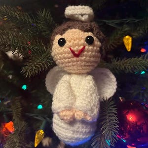 May include: A handmade crochet angel Christmas ornament. The angel has a cream-colored body, white wings, and a halo. The angel's hair is brown, and it has black eyes and a red smile. The ornament is hanging on a Christmas tree.