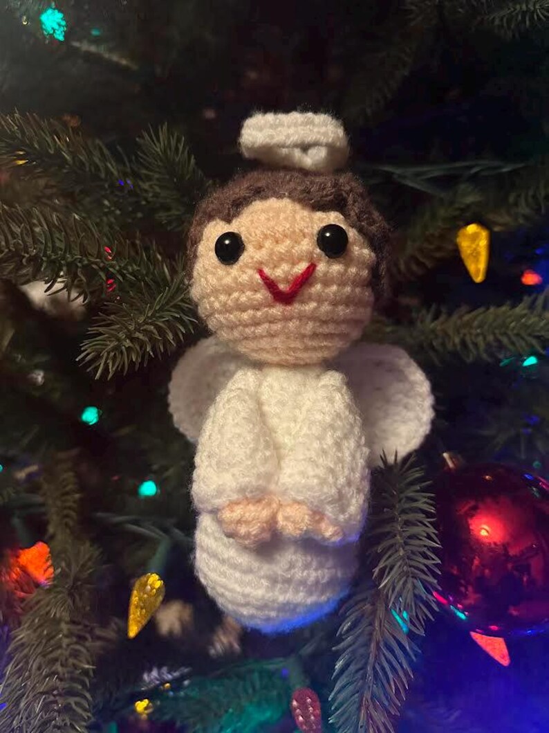 May include: A handmade crochet angel Christmas ornament. The angel has a cream-colored body, white wings, and a halo. The angel's hair is brown, and it has black eyes and a red smile. The ornament is hanging on a Christmas tree.