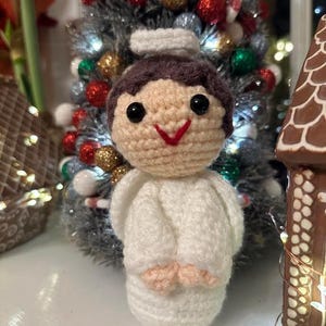May include: A handmade crochet angel ornament with a white robe, brown hair, and a red smile. The angel has black eyes and small peach-colored hands. A decorated Christmas tree is in the background.
