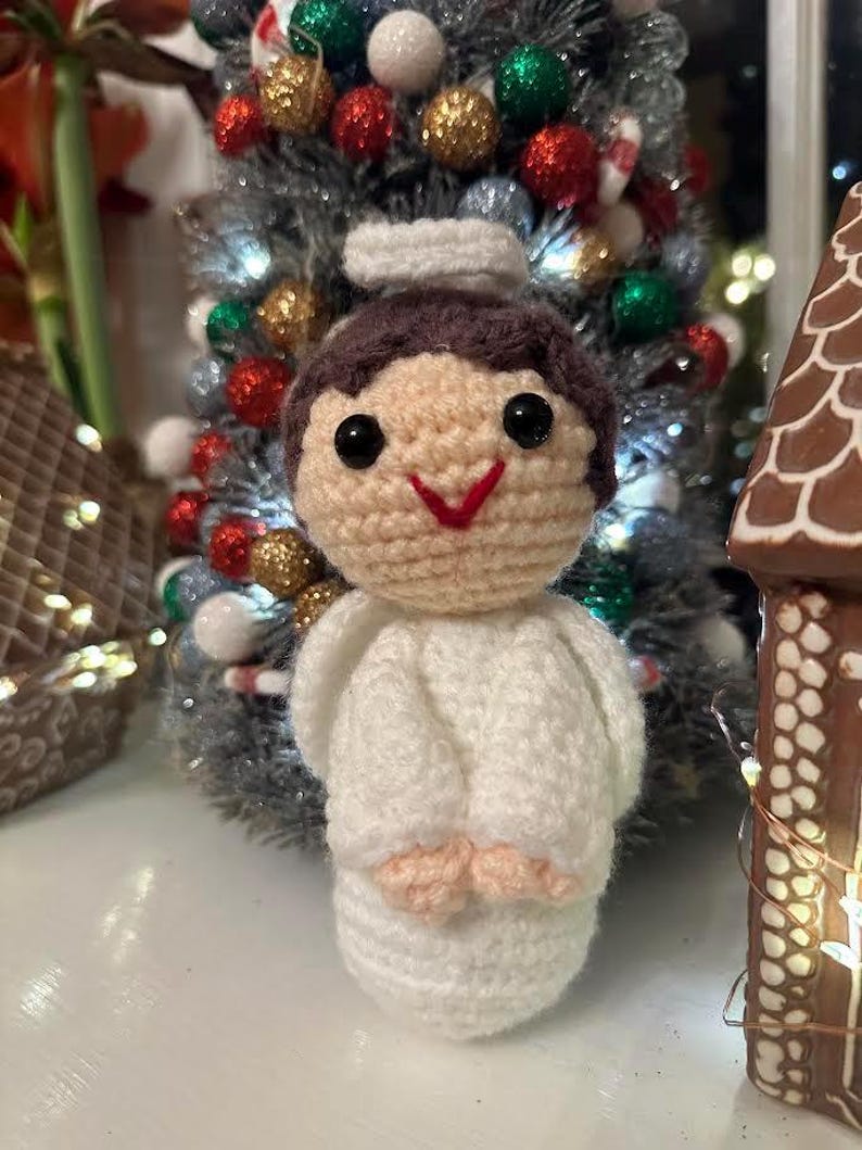 May include: A handmade crochet angel ornament with a white robe, brown hair, and a red smile. The angel has black eyes and small peach-colored hands. A decorated Christmas tree is in the background.