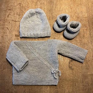 Op de afbeelding: Een lichtgrijze gebreide baby set, bestaande uit een muts, een overslag trui en een paar babyslofjes. De set is gemaakt van een zacht, getextureerd garen en is geplaatst op een houten oppervlak. De trui heeft striksluitingen.