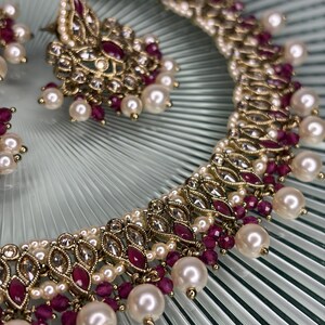 Puede incluir: Un conjunto de collar y pendientes de tono dorado ornamentado. El collar y los pendientes presentan una combinación de perlas de imitación, cristales transparentes y piedras preciosas de color rojo intenso. Las joyas se exhiben sobre una superficie azul claro acanalada.
