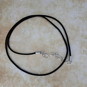 Peut inclure: Un collier en cordon noir doté d'un fermoir mousqueton argenté et d'une courte chaînette d'extension en métal argenté. Le cordon est lisse et circulaire, avec des embouts en métal argenté fixant le cordon au fermoir et à la chaînette.