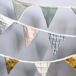 May include: A fabric bunting banner featuring multiple triangular flags in various patterns and colours, including sage green, white, pink stripes, blue stripes, and a mustard yellow botanical print with white animal illustrations, all attached to a white tape.