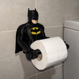 May include: A black Batman toilet roll holder. The holder is a figurine of Batman with a yellow bat symbol on the chest. It is mounted on a grey tiled wall and holds a roll of white toilet paper.