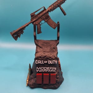 May include: A brown replica assault rifle displayed on a stand featuring the "Call of Duty: Modern Warfare" logo. The stand has three red rectangles and a bullet. The background is teal.