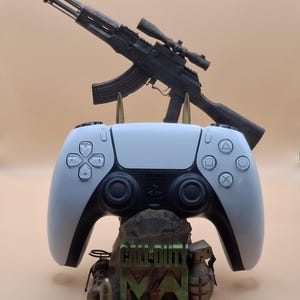 以下が含まれることがあります： 白と黒のPlayStation 5コントローラーが、緑色の「CALL OF DUTY MW」の文字が入った詳細なベースの上に置かれています。スコープ付きの黒いライフルがコントローラーの上に配置されています。ベースには手榴弾と弾丸の詳細が含まれています。