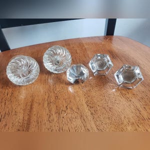 May include: A collection of clear glass knobs and pulls. Two are round with a spiral design, and three are hexagonal. The hardware is displayed on a wooden surface.