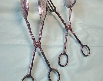May include: Three antique silver serving utensils: a spoon, a fork, and a spatula-like tool. Each utensil has a scissor-like handle with a decorative center. The spoon and spatula have rounded heads, while the fork has three tines. The utensils are arranged on a light green surface.