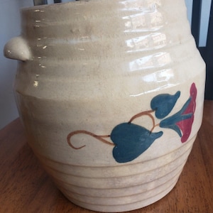 May include: Cream-coloured ceramic pot with a floral design. The pot has a wide, ribbed body and a small handle on the side. The floral design features blue leaves and a red flower. The pot is likely used for decorative purposes.